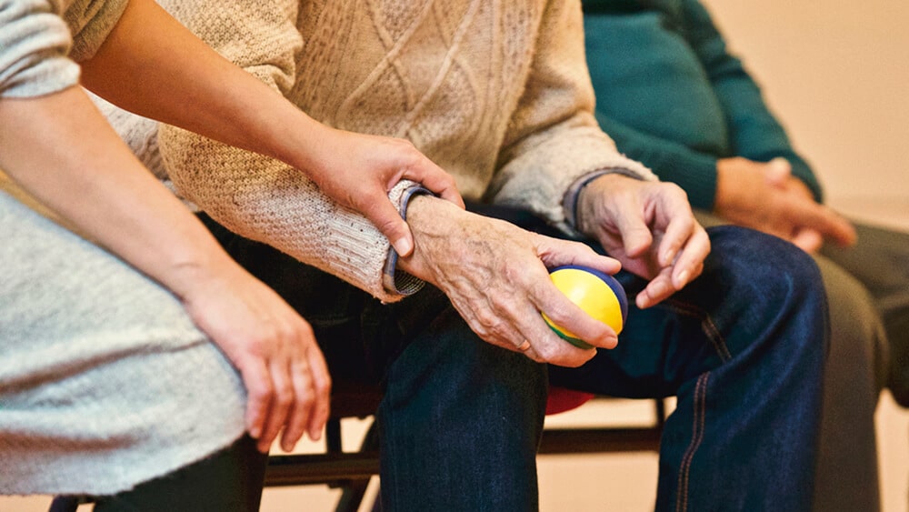 Elder person sitting down holding ball in hand, person sitting next to them is touching their wrist.